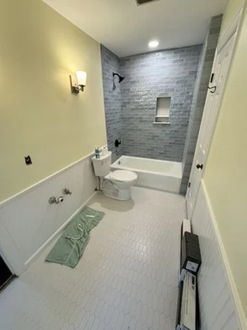 A small bathroom with light yellow walls, a white toilet, a bathtub with gray tiles, a built-in shelf, a wall-mounted light fixture, and a green bath mat on a white tiled floor.