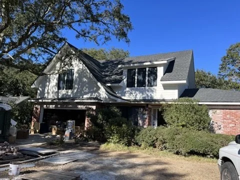 Two-story house with white siding and a steep roof, surrounded by trees and bushes, under a clear blue sky.