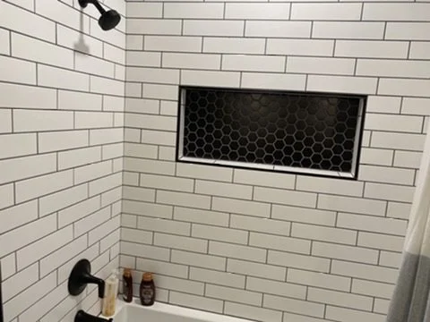 A shower with white subway tiles, a black showerhead, and two bottles in the corner, with a black honeycomb-patterned window.
