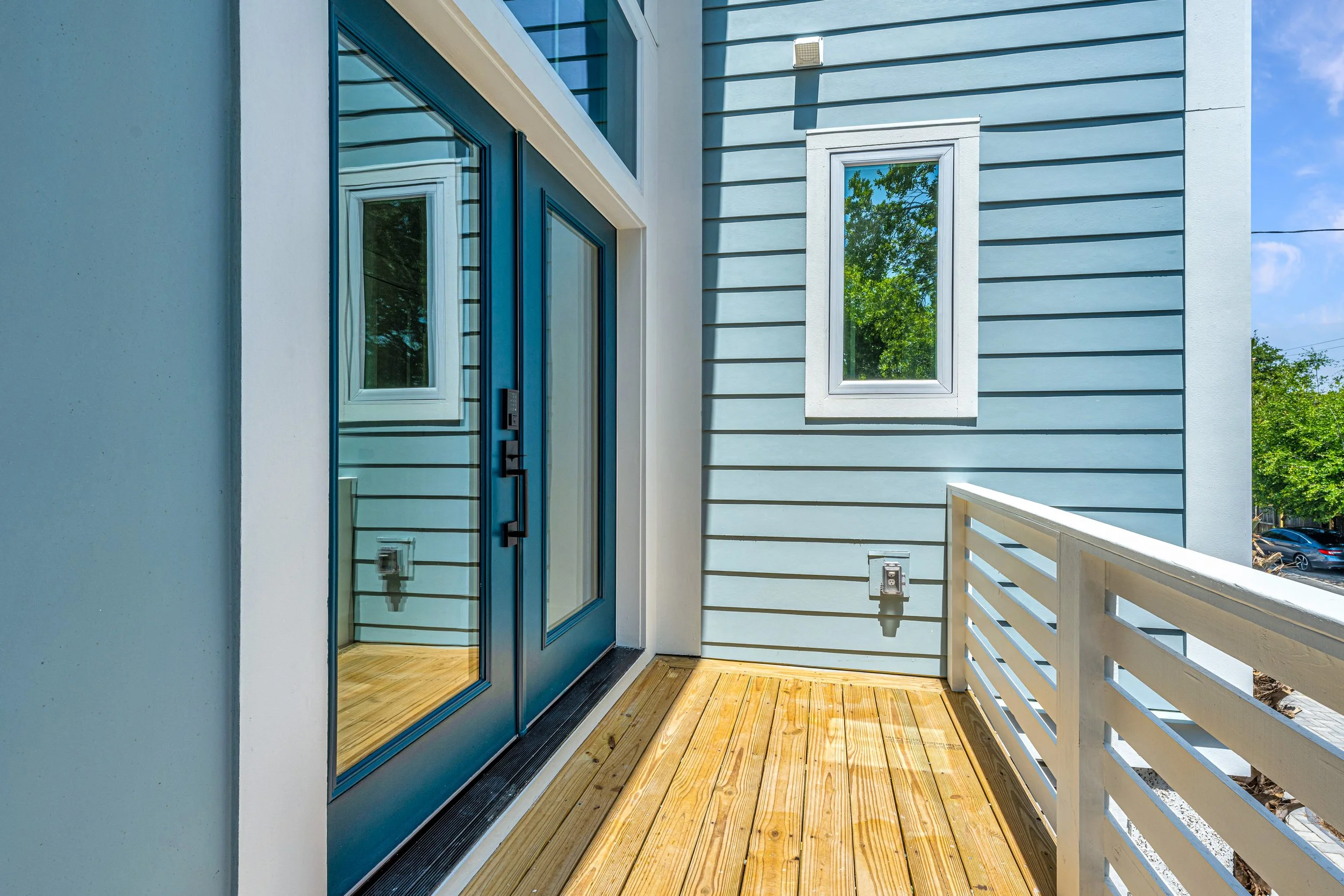 View of a small outdoor balcony with a new wooden deck, blue double glass doors, a white railing, and a light blue house wall with a small window.