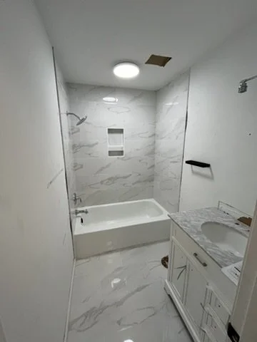Bathroom with bathtub, marble-like walls and floors, white vanity with sink, mirror, and some construction damage on the ceiling.