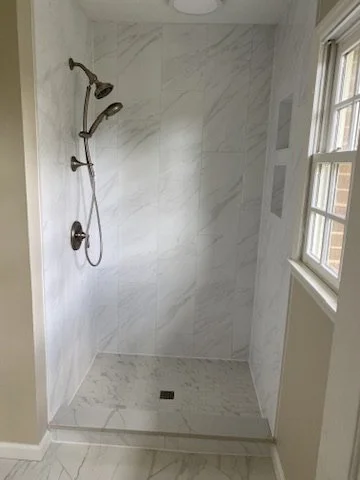 Empty walk-in shower with marble walls and floor, a handheld showerhead, a fixed showerhead, and two windows in a bathroom.