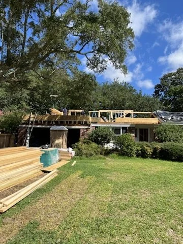 House under construction with wooden beams and construction materials outside, large tree and blue sky with clouds