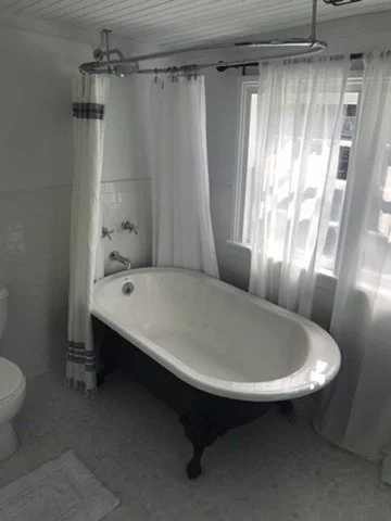 A vintage clawfoot bathtub with a white interior and dark exterior, located in a bathroom with white curtains and some natural light coming through the window.