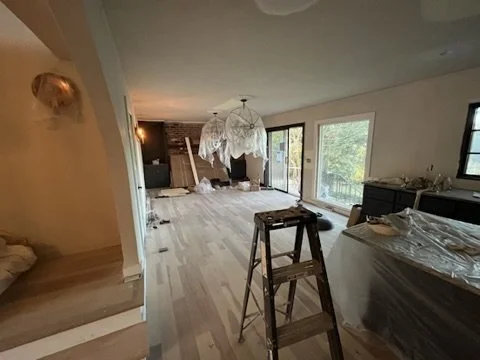 Interior of a house under renovation, with a ladder, plastic-covered furniture, a chandelier, and sliding glass doors leading outside.