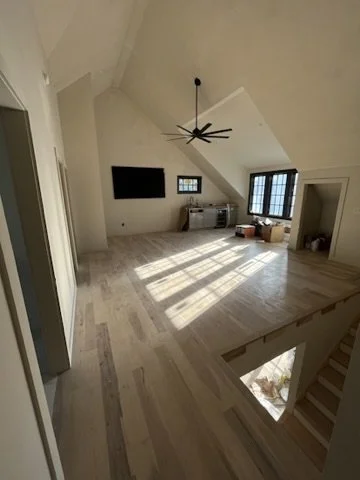 A spacious, empty living room with high vaulted ceilings, light-colored wood floors, large windows letting in sunlight, a modern black ceiling fan, a wall-mounted flat-screen TV, and a small balcony or stairwell opening.