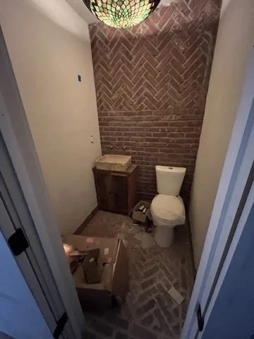 Small bathroom with brick wall, toilet, and a box on the floor holding tools or supplies, with a decorative hanging lamp above.
