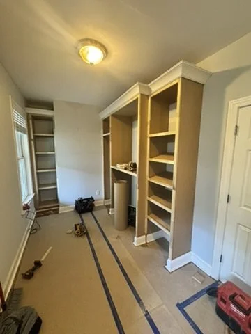 Empty room with built-in shelves, a window with blinds, and a closed door, illuminated by a ceiling light.