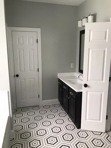 Bathroom with a closed door, a vanity with a mirror and a black cabinet, and a white folding door, mosaic tile flooring with black and white geometric patterns.