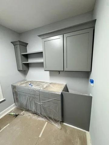 Gray kitchen cabinets and shelves in a small room, with a covered appliance on the floor and paint supplies nearby.