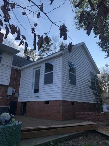 A house with a brick foundation and white siding, with three windows on the side visible, trees overhead, and the sky visible.