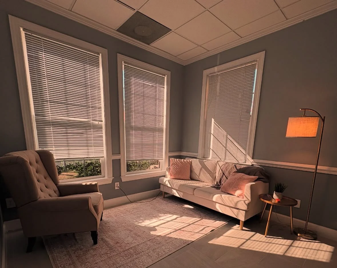 Living room with three large windows with closed blinds, a beige armchair, a white sofa with pink and dark pillows, a small wooden side table with a plant and candles, and a tall floor lamp with a beige lampshade, illuminated by sunlight casting shadows through blinds.