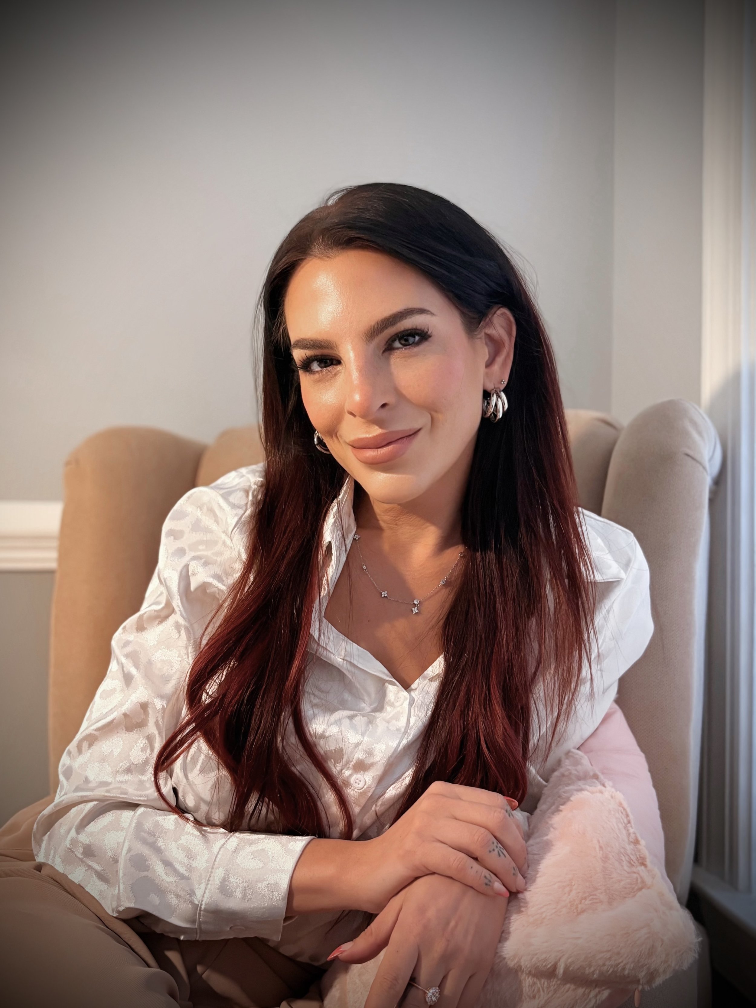 A woman with long, dark hair and light makeup sitting on a beige armchair, smiling softly, wearing a white patterned blouse, silver jewelry, and holding a pink fluffy pillow.