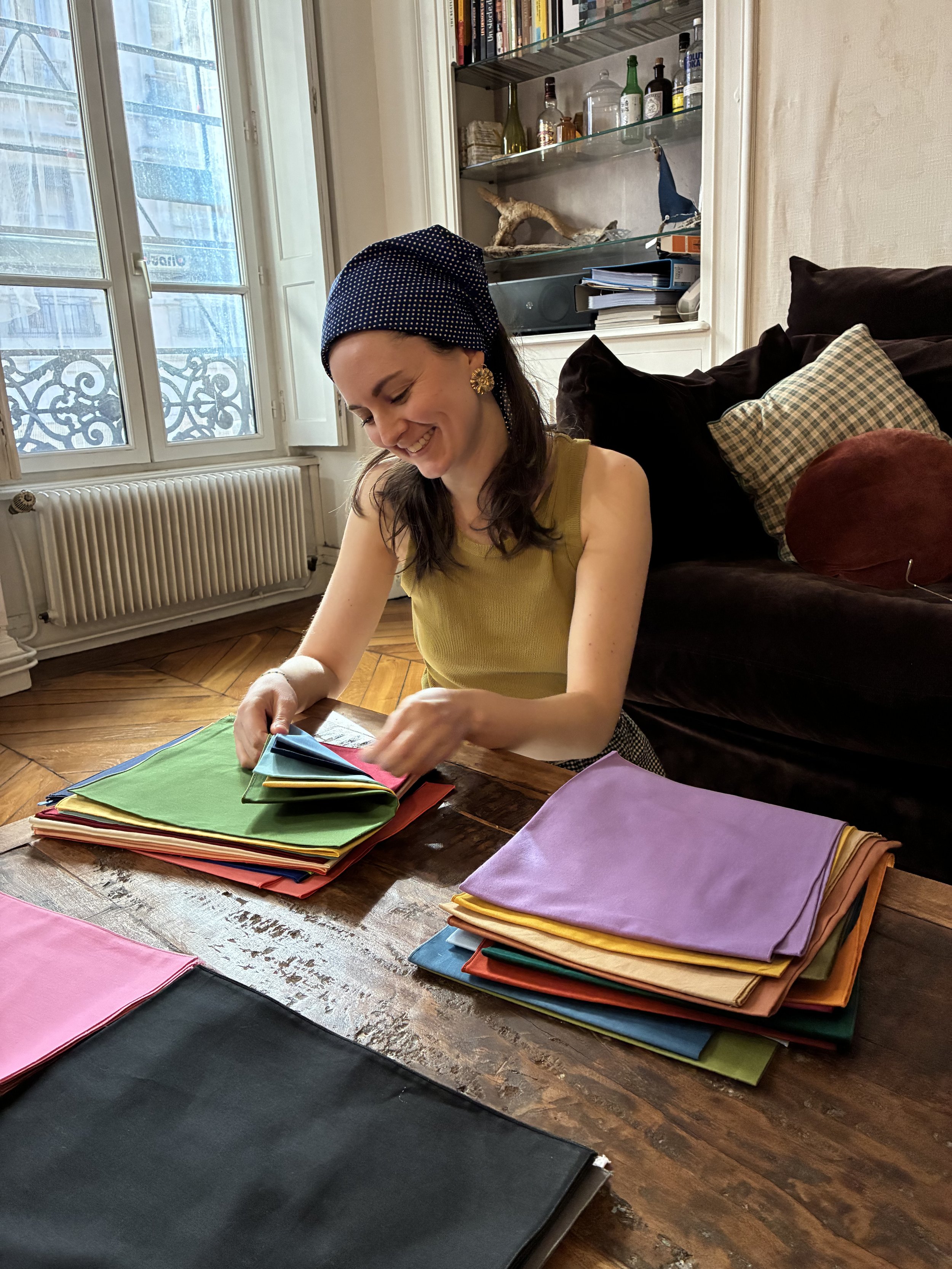 Une femme souriante et habillée en jaune, portant un foulard bleu à pois, assise à une table en bois, triant des morceaux de tissu colorés.