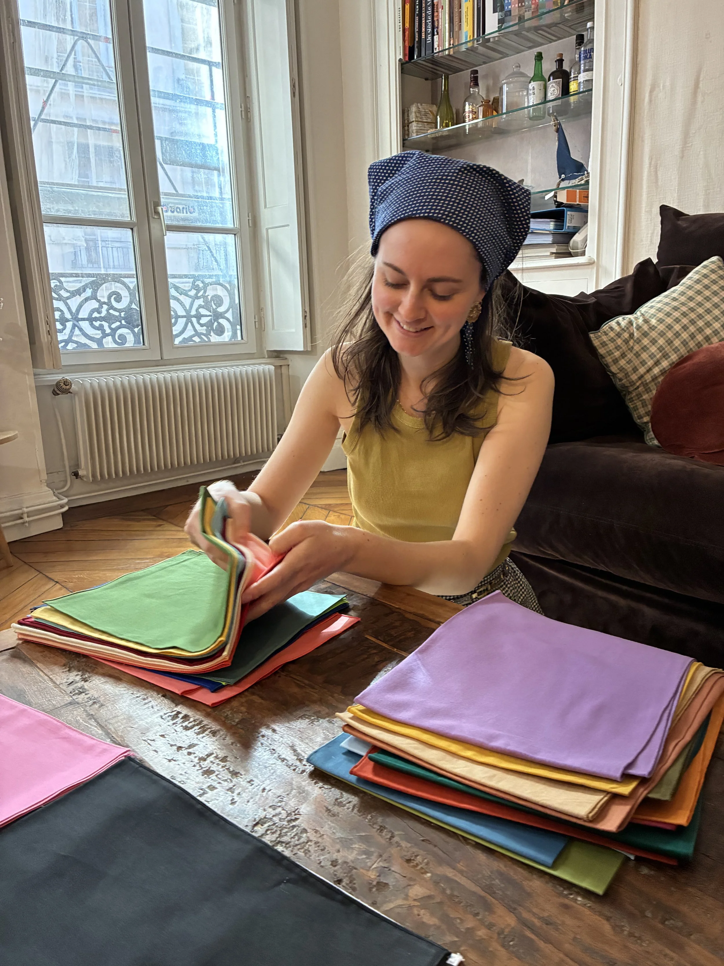 Une femme souriante en train de trier des tissus colorés sur une table dans une pièce lumineuse, avec un large window et une bibliothèque en arrière-plan.