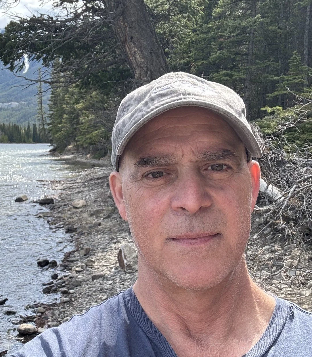 A man taking a selfie outdoors near a lake with trees and mountains in the background.