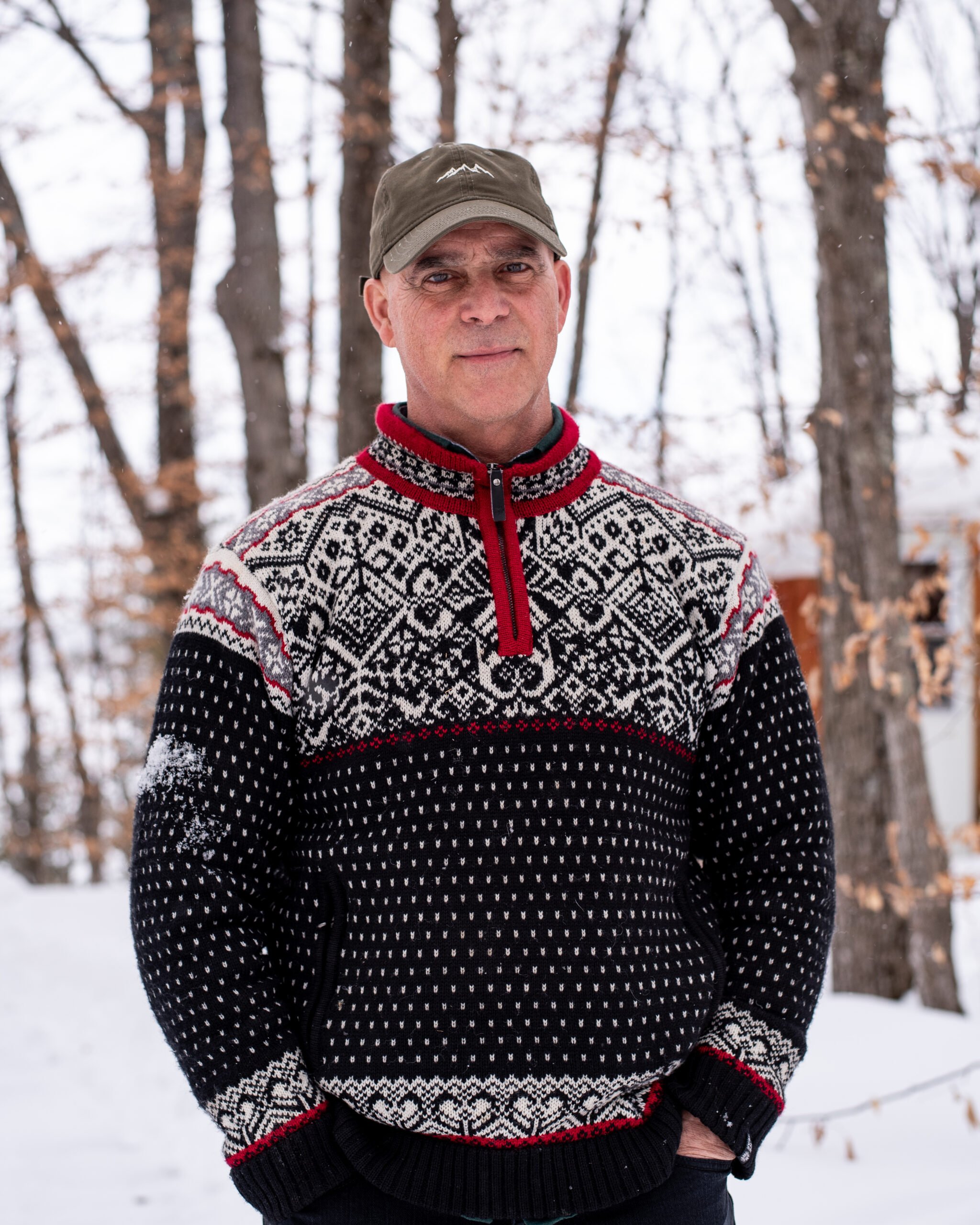 A middle-aged man wearing a patterned knit sweater and a baseball cap posing outdoors in a snowy forest during winter.