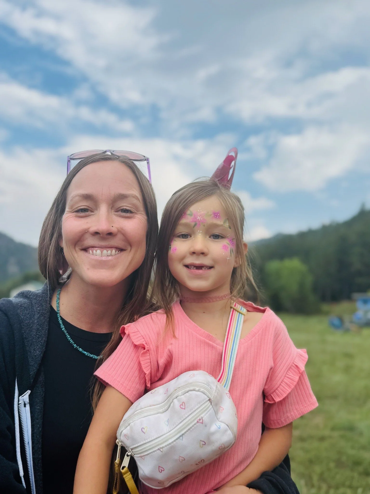 A woman and a young girl taking a selfie outdoors with a blue sky, clouds, and green hills in the background. The girl has face paint and is wearing a pink shirt, while the woman has sunglasses on her head and a smile.