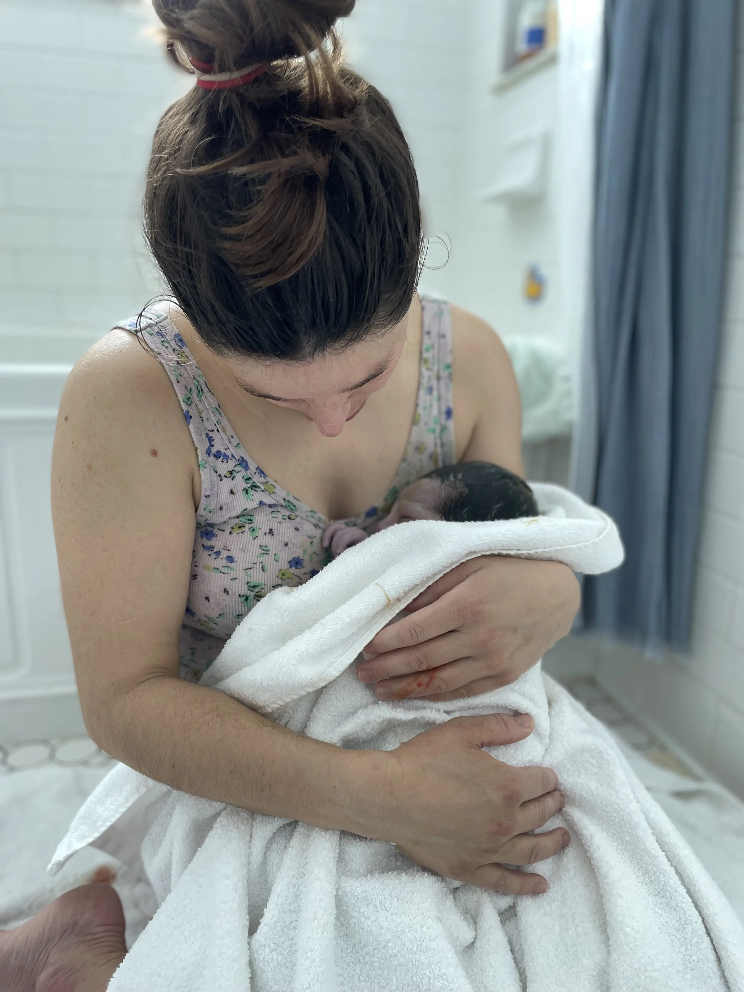 Woman holding a newborn baby wrapped in a white towel, looking at the baby in a room.
