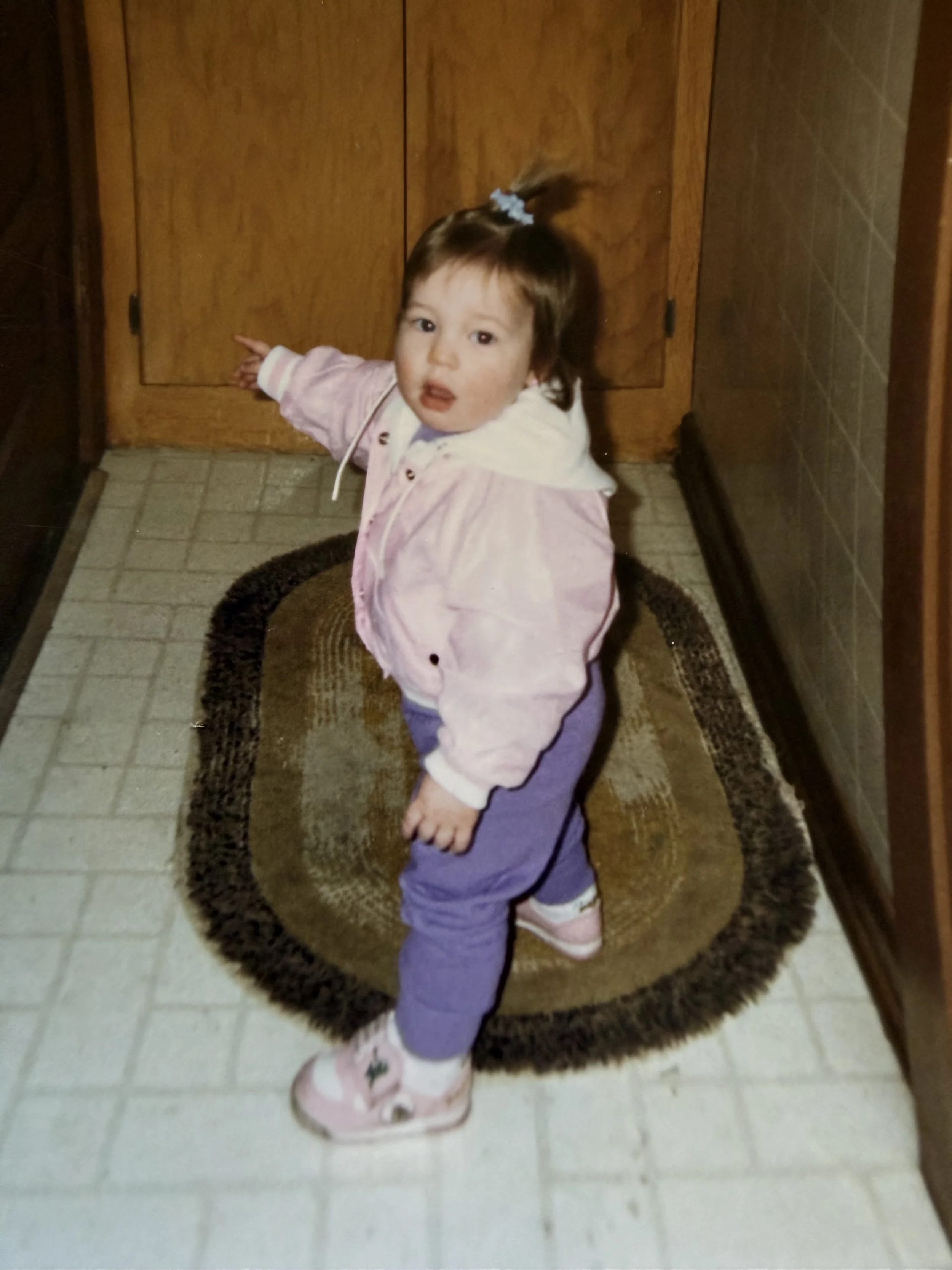 A young girl with brown hair tied up with a blue hair tie, wearing a pink jacket, purple pants, and pink shoes, standing on a brown rug in a kitchen, touching cabinet doors with a surprised or curious expression.