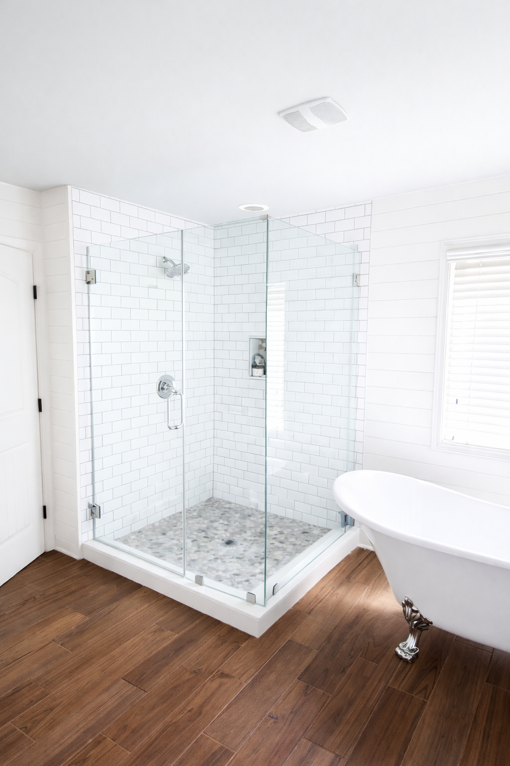 Custom bathroom remodel featuring a glass-enclosed shower with white subway tile and pebble shower floor, white clawfoot bathtub, and warm wood flooring by Wilken Precision Remodeling.