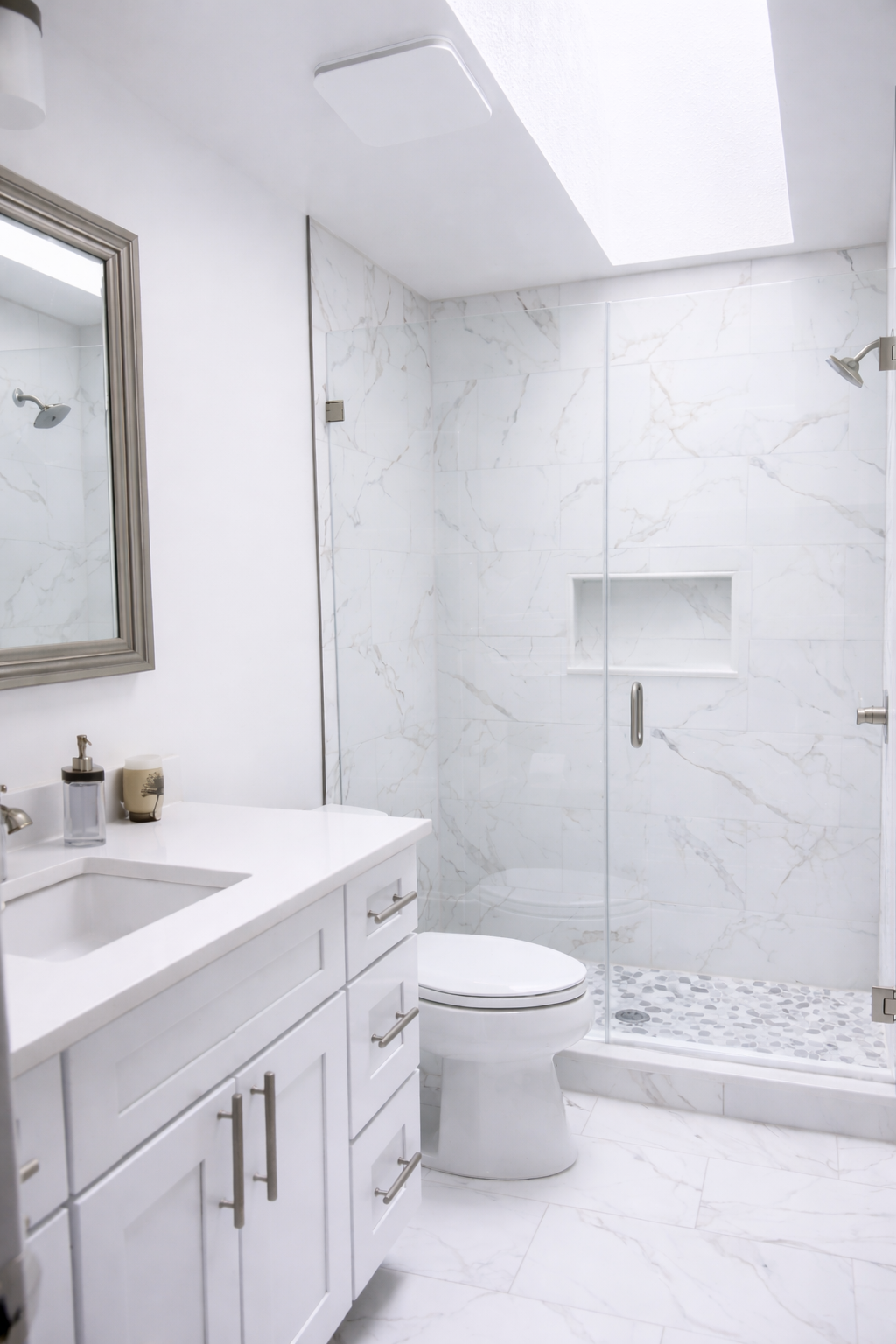 Modern custom bathroom remodel featuring white marble tile walls and floors, frames glass shower enclosure, white vanity, and clean minimalist finishes by Wilken Precision Remodeling.