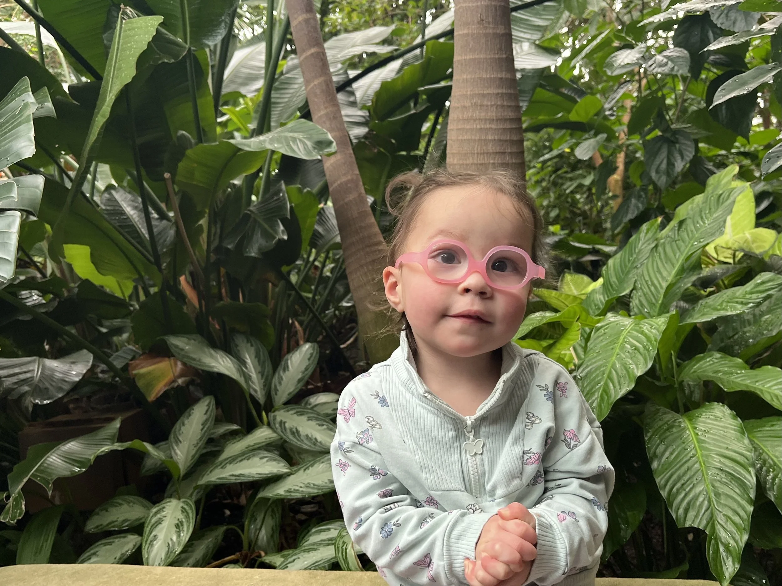 Girl with FOXP1 syndrome in pink glasses and a pale turquoise jacket, posing with clasped hands in front of some trees
