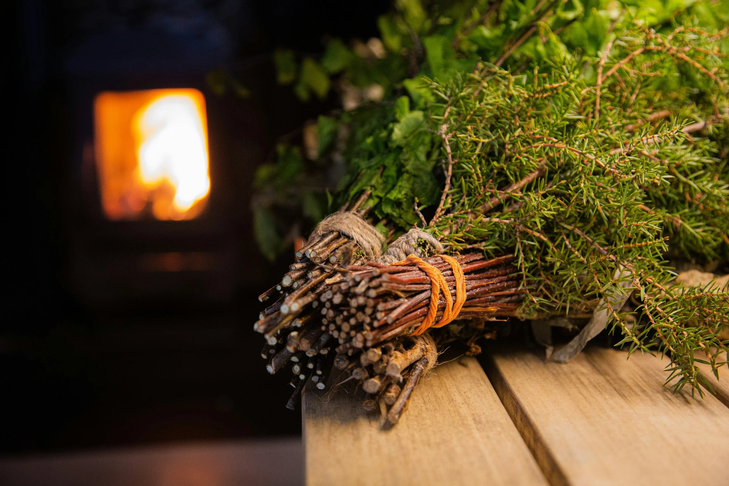 A bundle of sticks and green foliage tied with twine and orange rubber bands, resting on a wooden surface, with a glowing fire in the background.