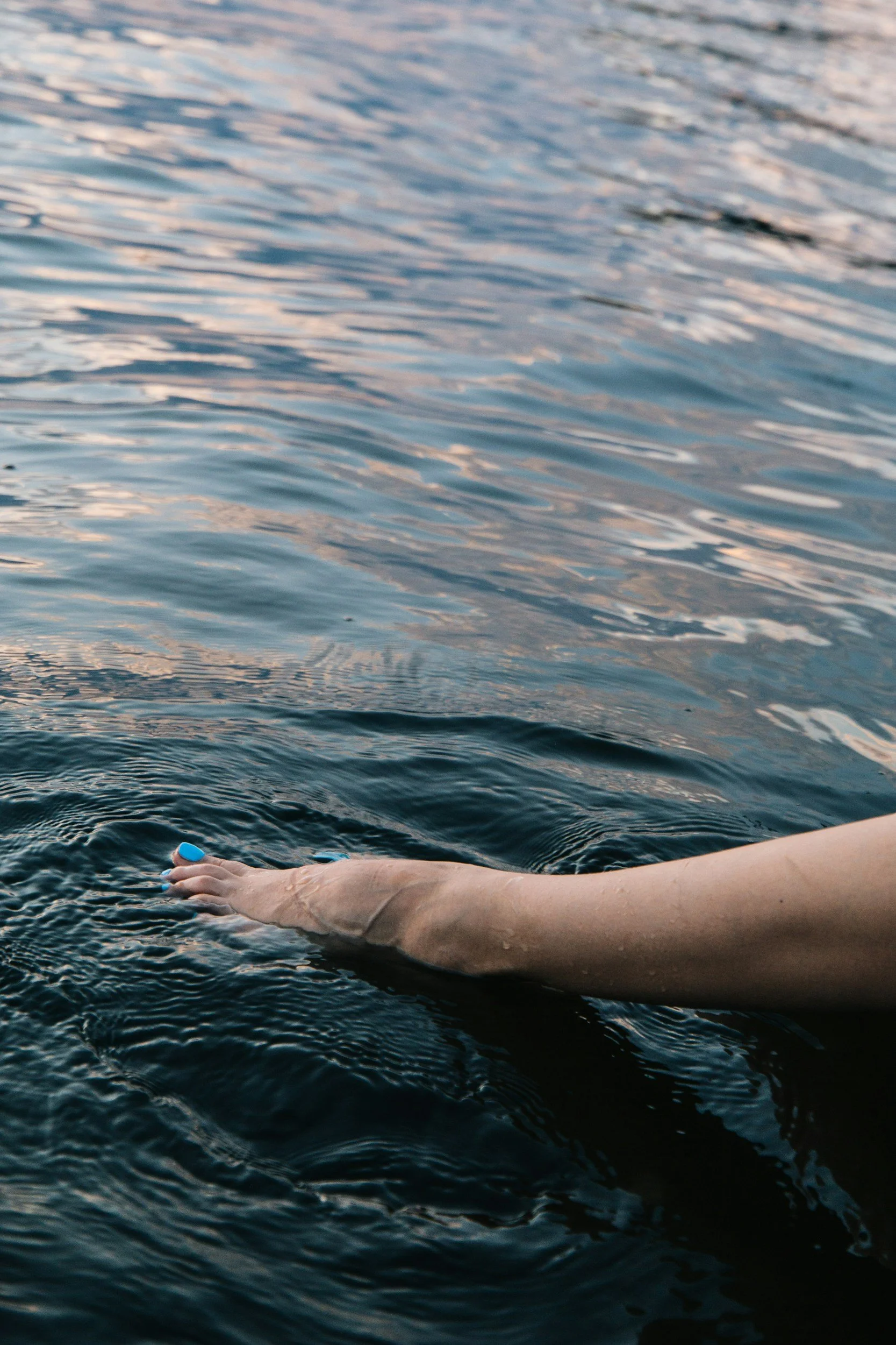 A person's arm and hand with blue-painted nails extending into dark water, creating ripples.