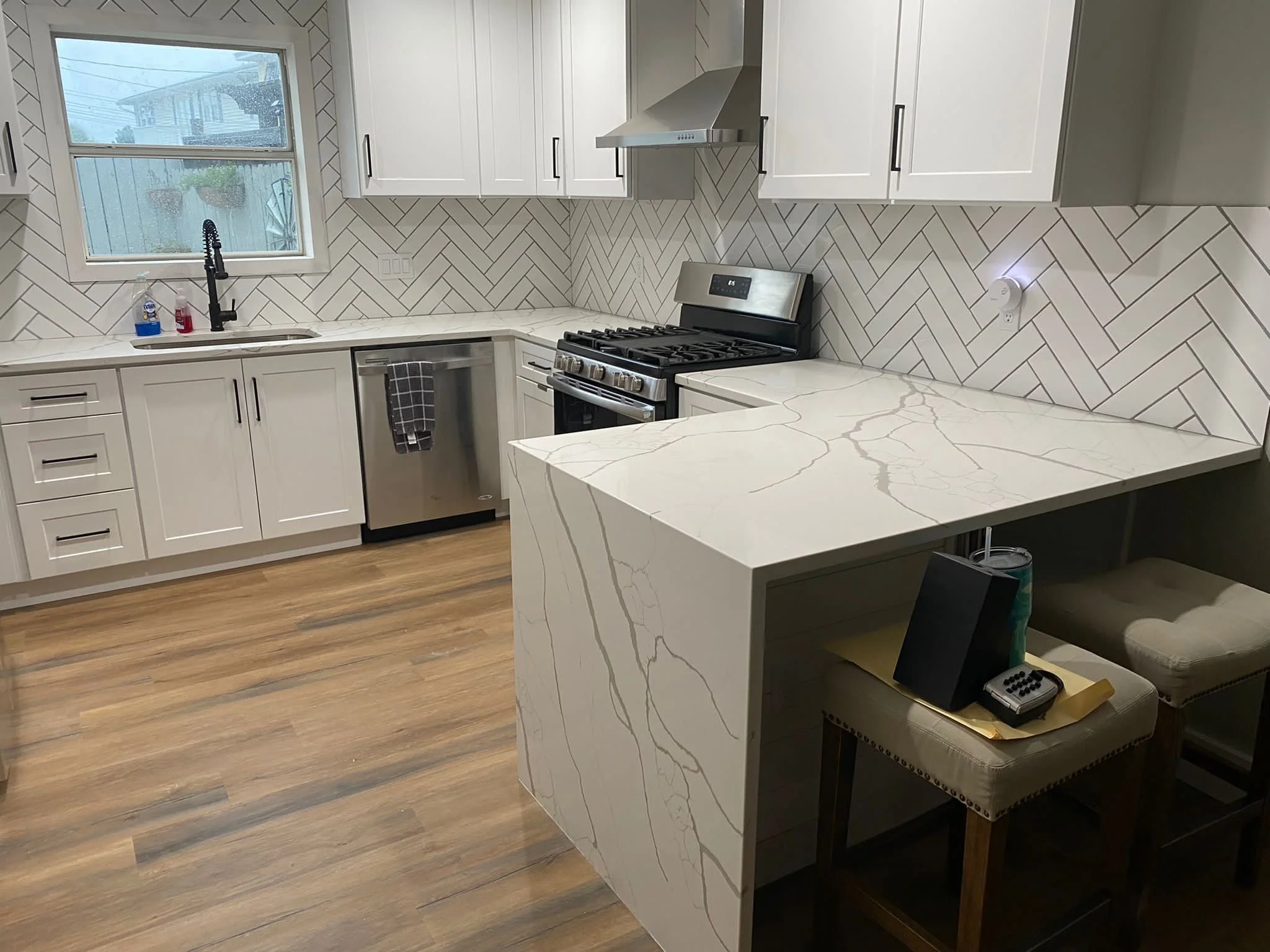 Modern kitchen with white cabinets, marble countertops, a gas stove, stainless steel dishwasher, and herringbone-patterned tile backsplash; hardwood flooring, a window with plants outside, and a small seating area with chairs and a tray holding books and a phone.