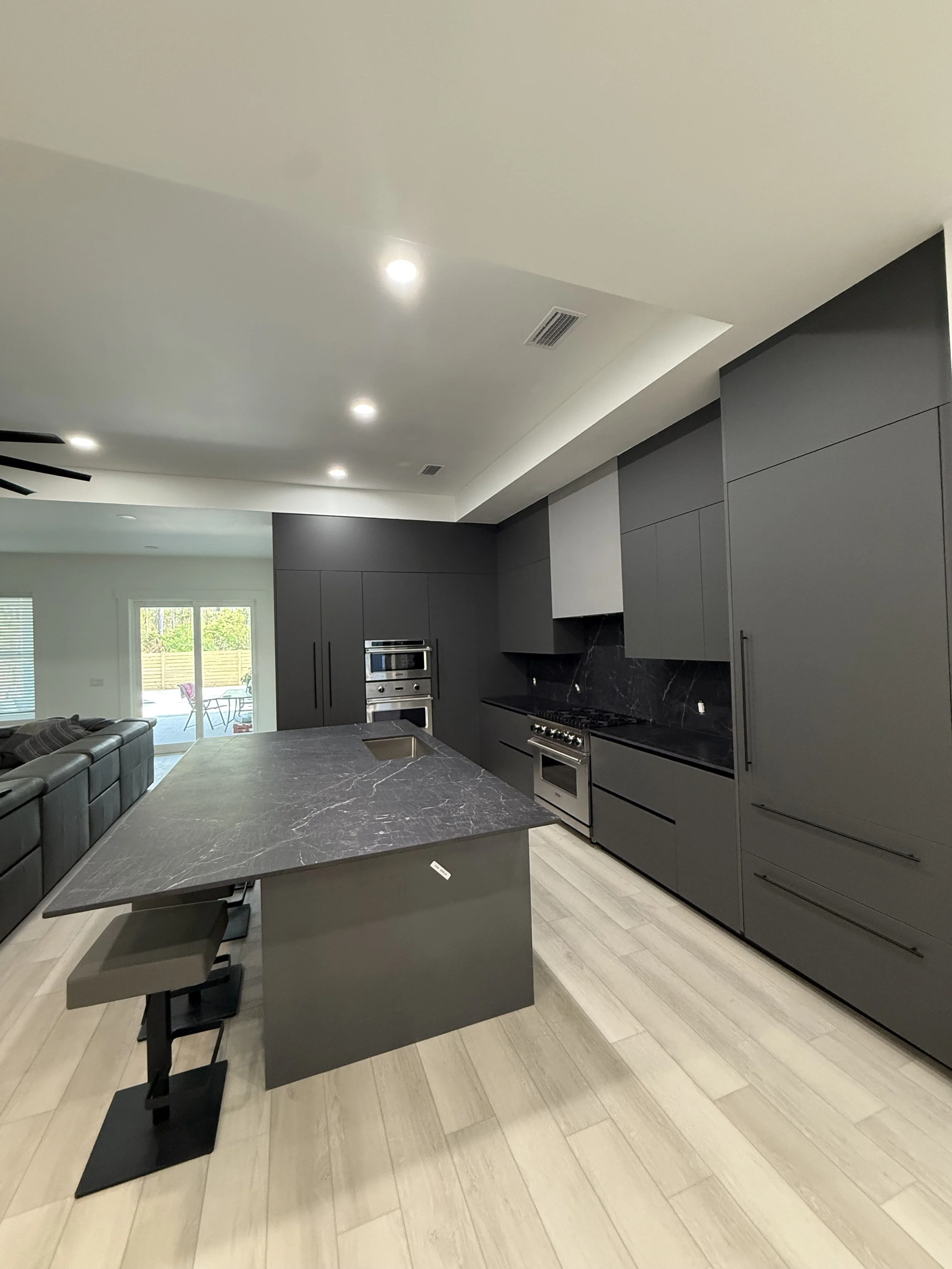 Modern kitchen with dark gray cabinets, black marble countertops, an island with a matching marble top, stainless steel appliances, light wood flooring, and a sliding glass door leading outside.
