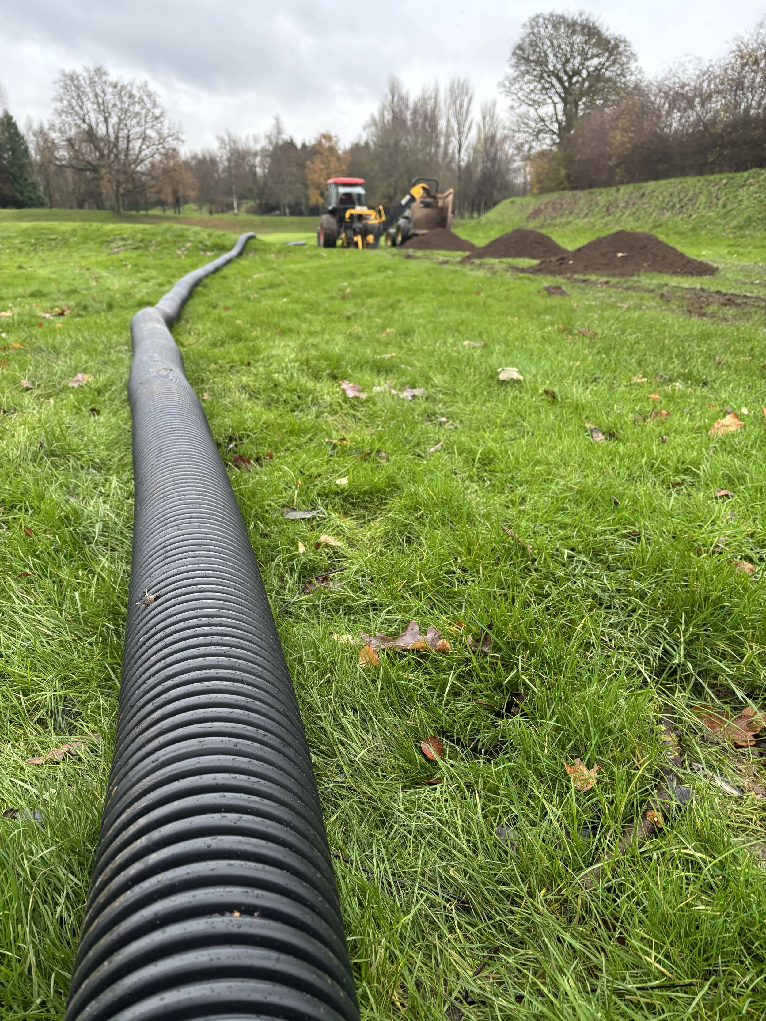 A large black drainage pipe on a grassy field leading toward a backhoe digging a trench, with piles of dirt nearby.