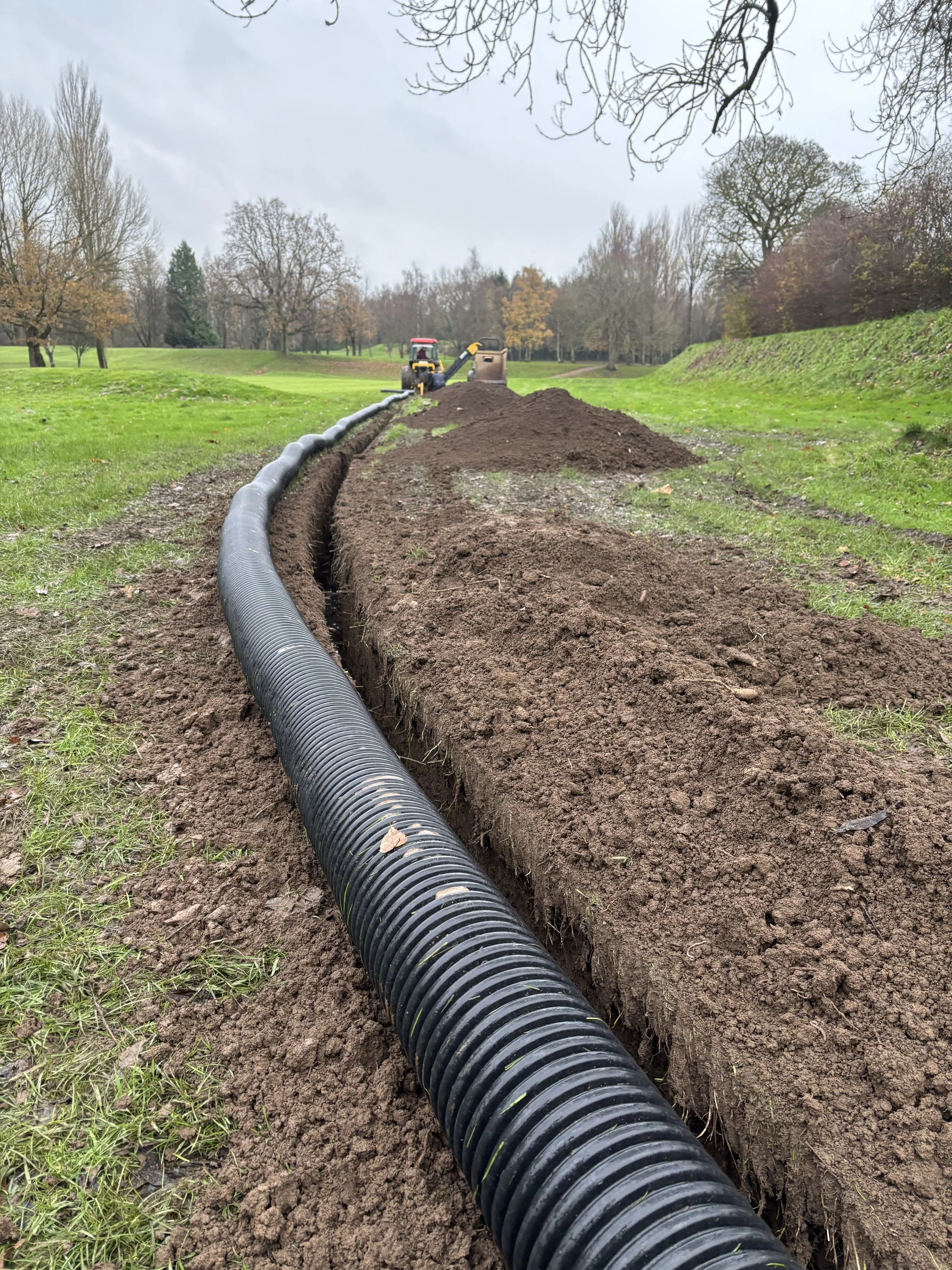 A large black flexible pipe running through a trench in a grassy field, with a tractor in the distance, on a cloudy day.