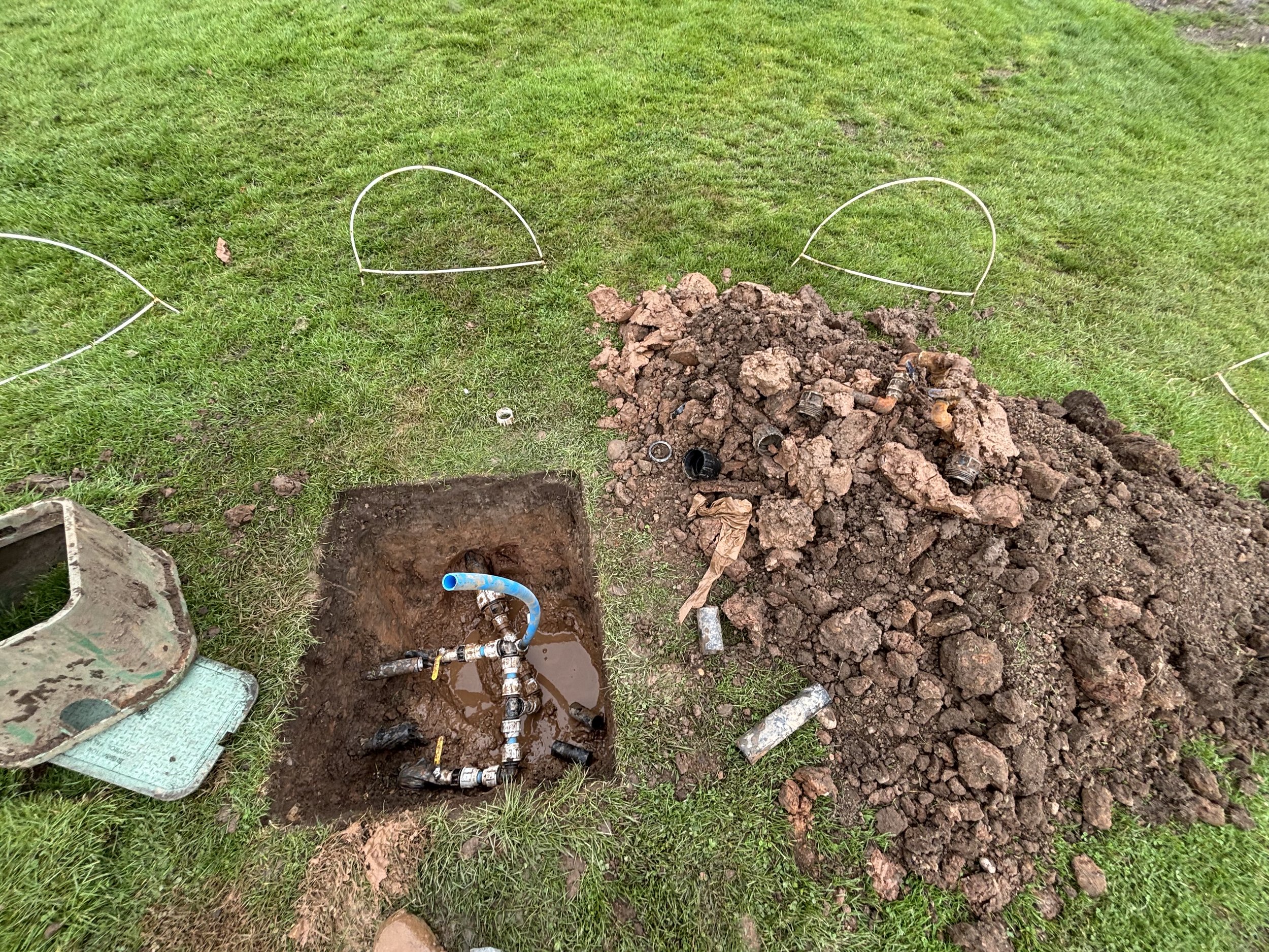 A repair site with an underground water pipe connected by various fittings in a dug hole, beside a large pile of dirt and a small fenced-off area in a grassy yard.