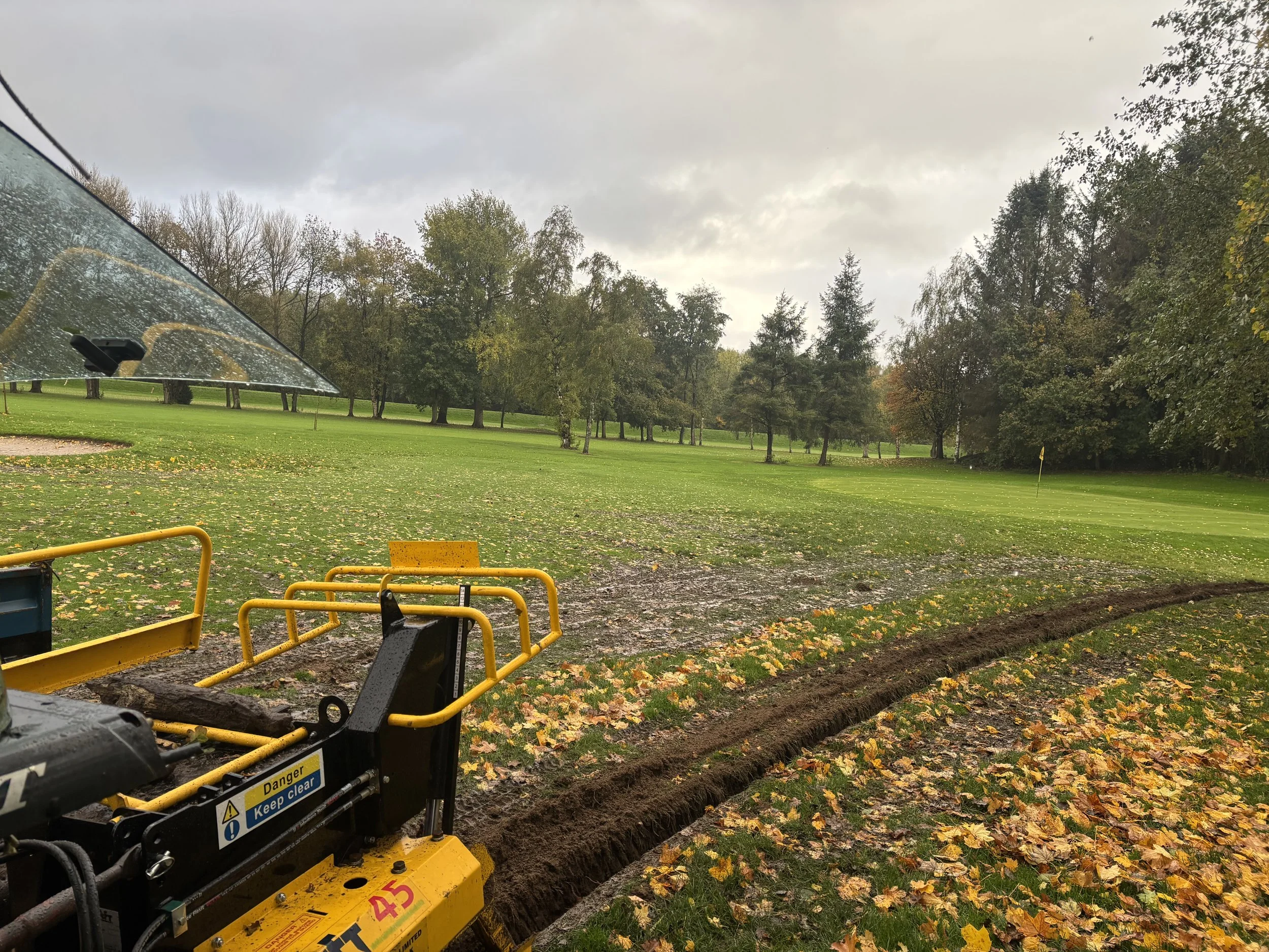 A golf course under cloudy skies, with construction equipment digging a trench in the foreground, fallen autumn leaves scattered across the grass, and a flagstick on the green in the distance.