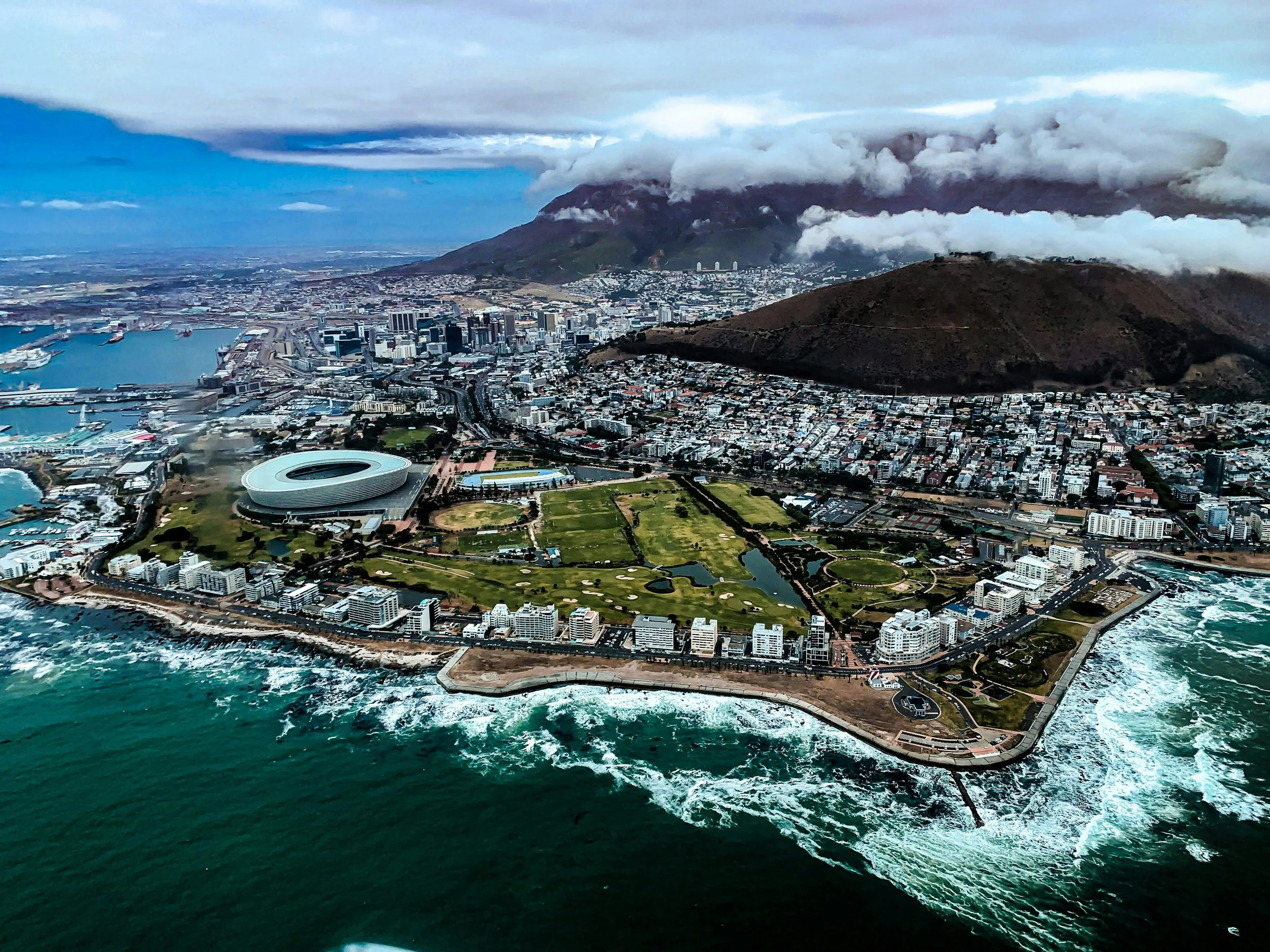 Aerial view of a city with a stadium, green parks, water, mountains, and cloudy sky.