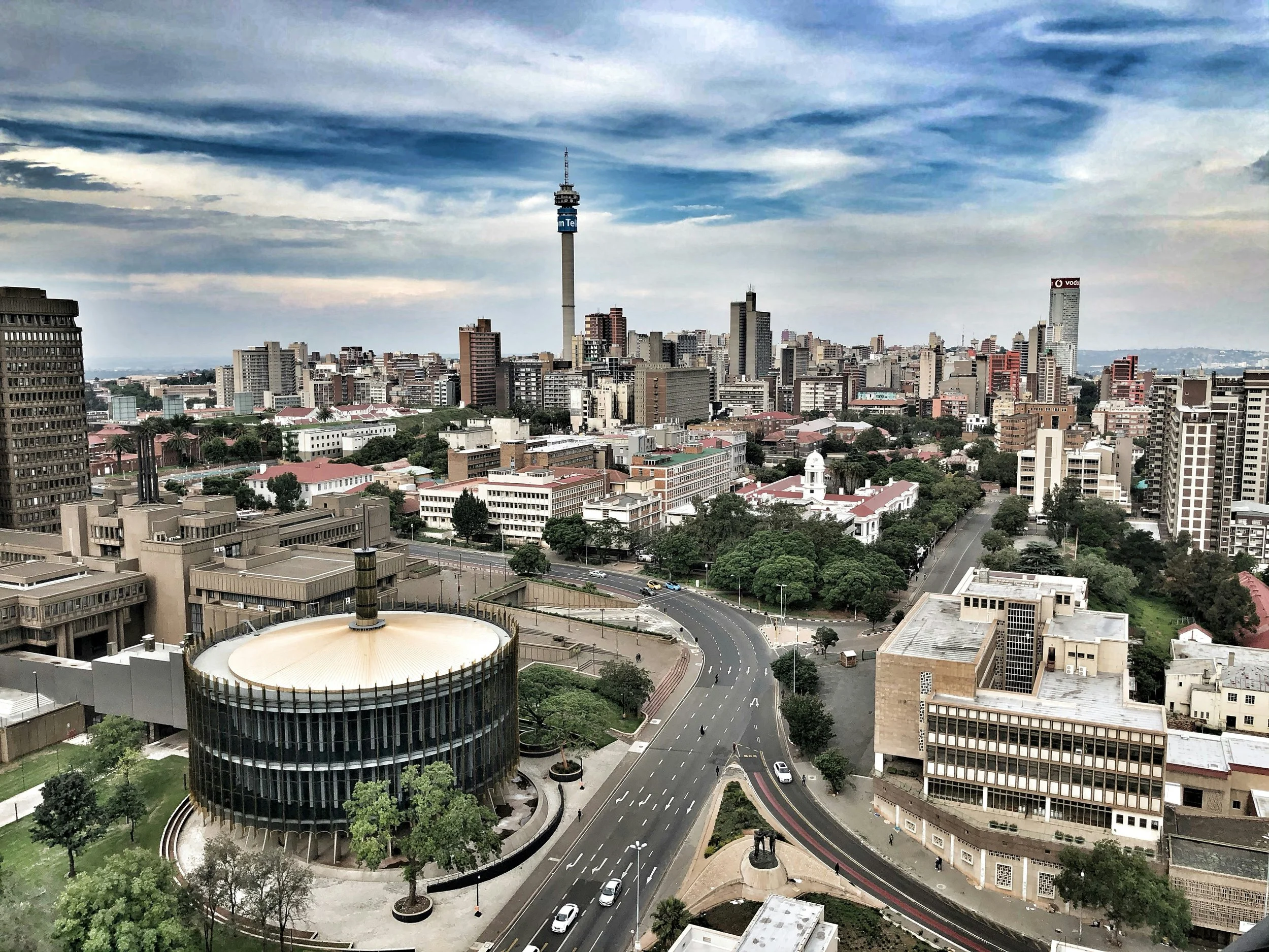 A cityscape of Johannesburg with tall buildings, a round building with a golden roof, and a prominent telecommunications tower under a partly cloudy sky.