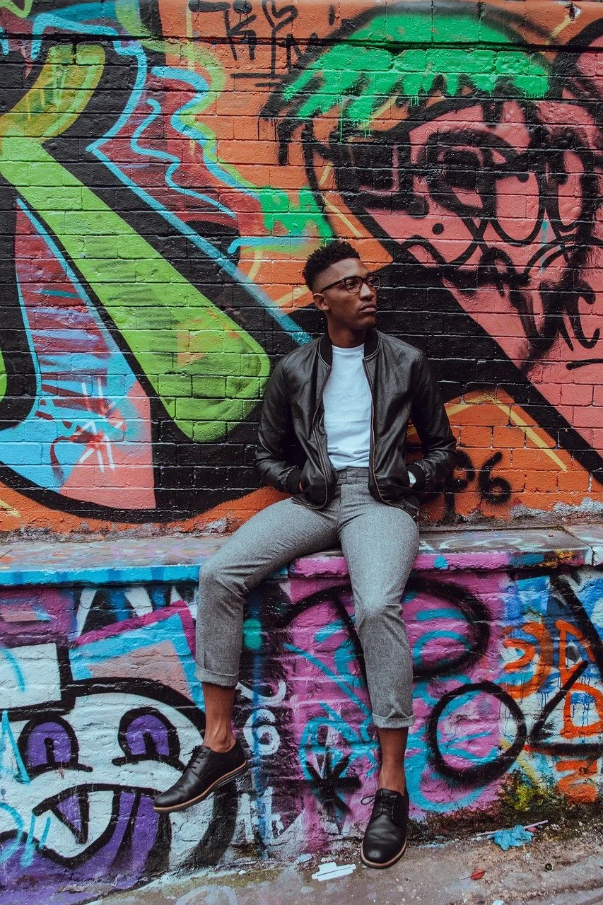 A young man sitting on a graffiti-covered ledge against a colorful graffiti wall, wearing a black leather jacket, white shirt, gray pants, and black shoes, with glasses and short hair.