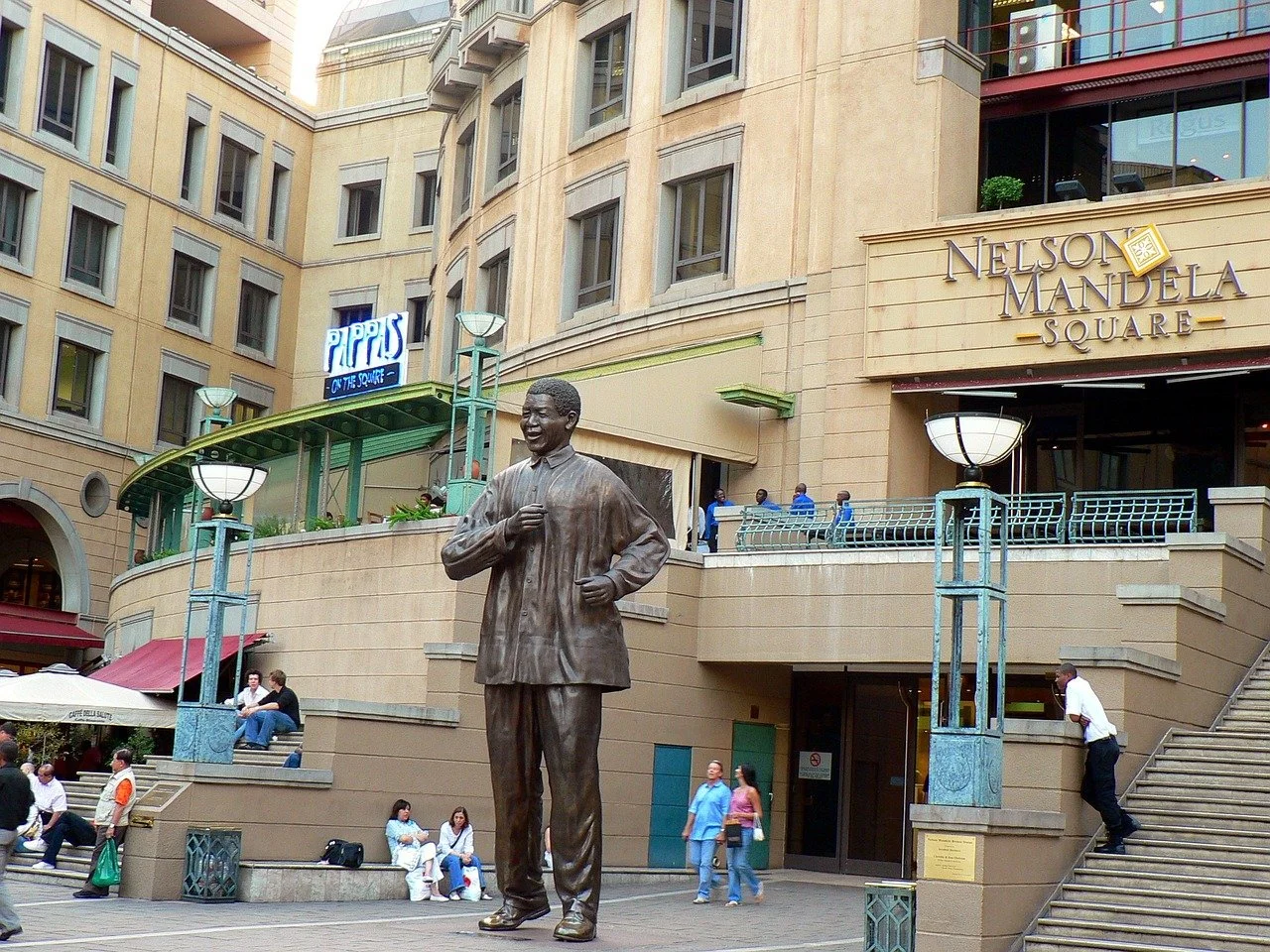 Statue of Nelson Mandela in Nelson Mandela Square at an outdoor shopping area with people sitting, walking, and walking up stairs. Building in the background has signs including one for 'Pipps on the Square'.
