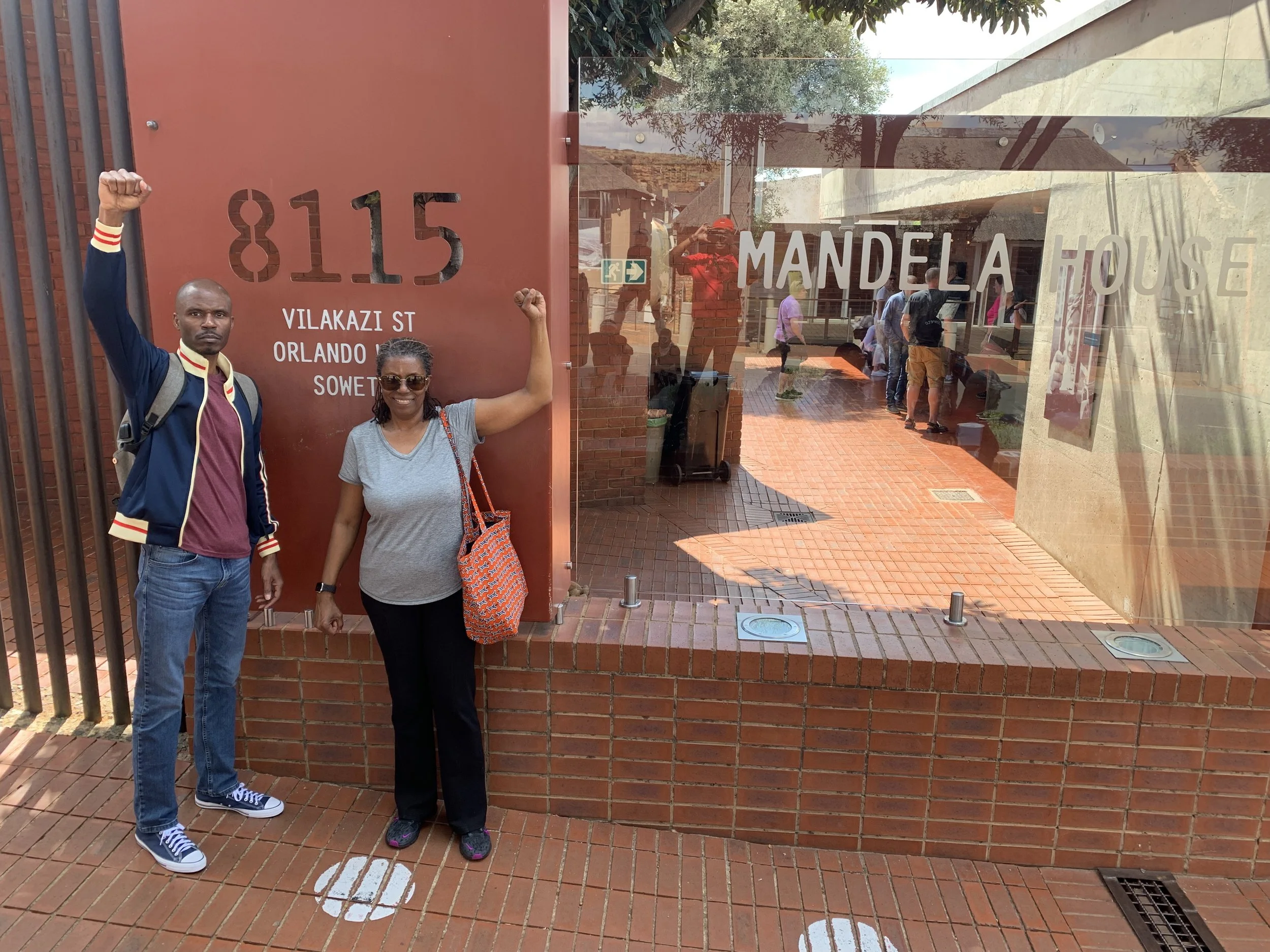A man and a woman standing outside the Mandela House, with the man flexing his arm and the woman smiling and pointing upward. The Mandela House sign and address are visible in the background.