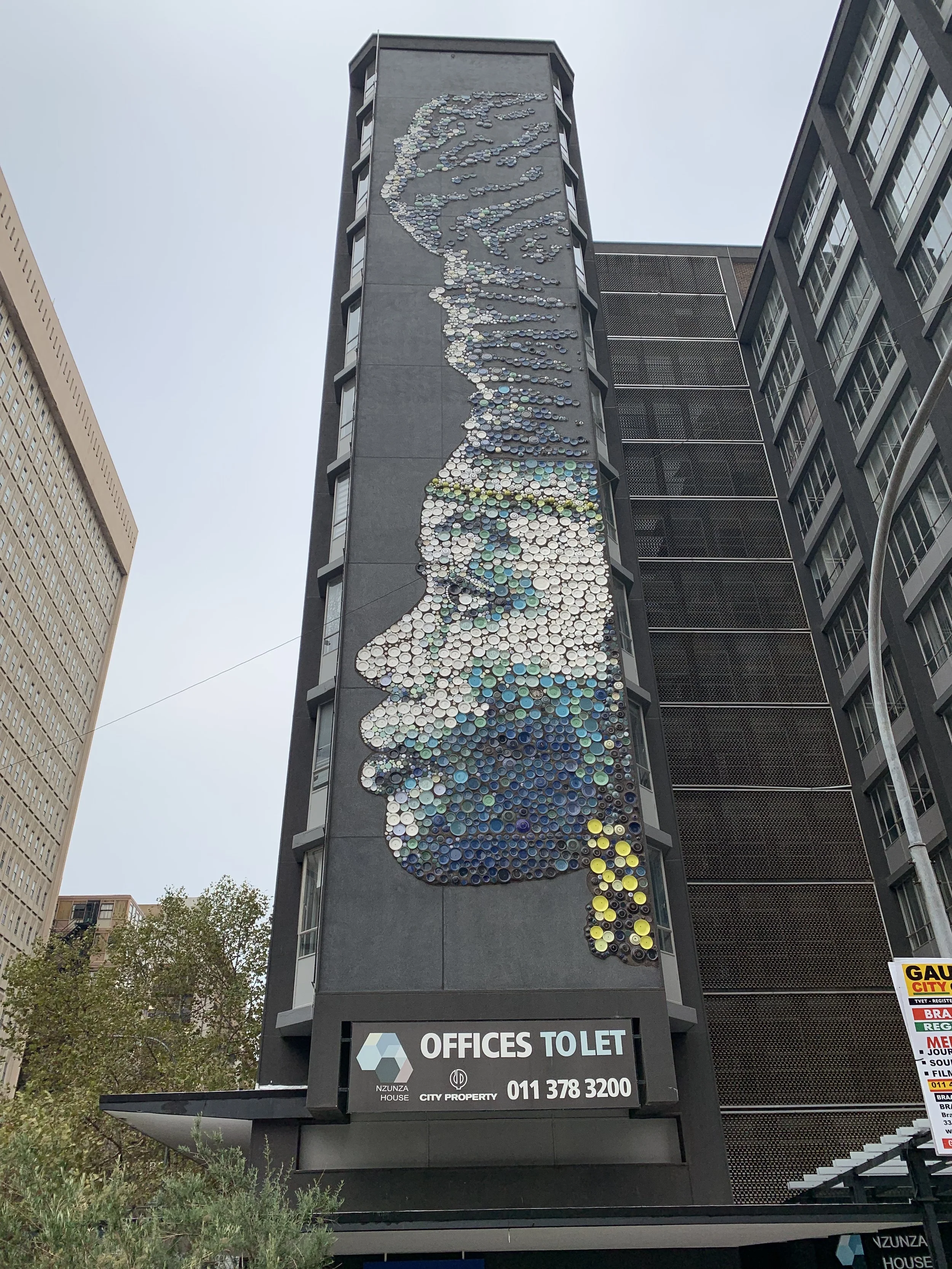 Tall building with a large mosaic portrait made of circular tiles on its facade, depicting a woman with curly hair. A sign at the bottom advertises office space for rent.