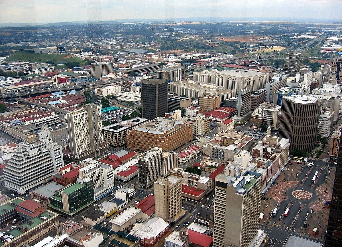 Aerial view of a city with high-rise buildings, roads, and a circular plaza with buses and people, with a landscape of hills and fields in the background.