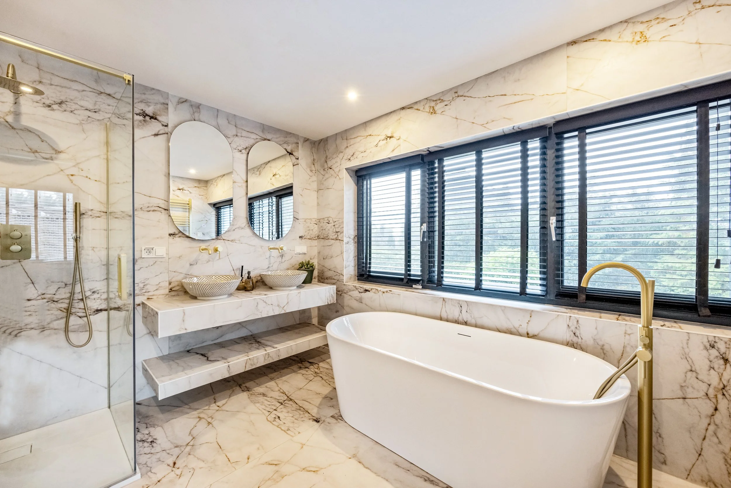 Luxe badkamer met marmeren muur en vloer, bad, dubbele wastafel met ovalen spiegels, en grote ramen met zwarte jaloezieën.