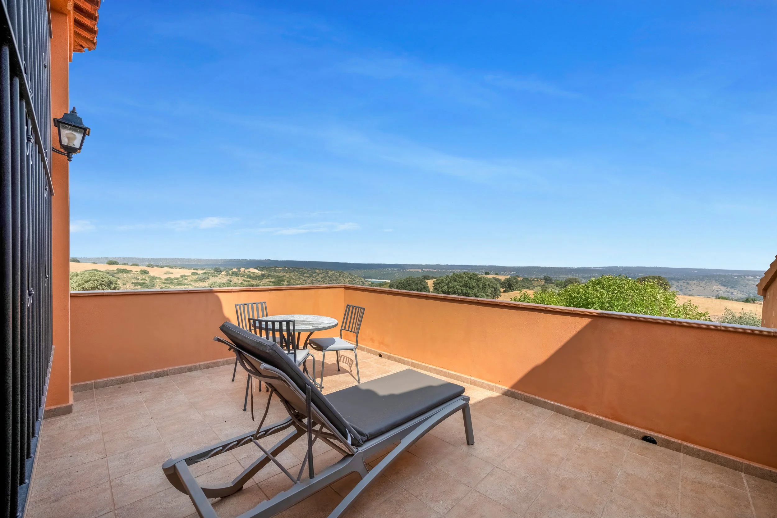 Een zonnig balkon met een ligstoel, een ronde tafel met vier stoelen, omgeven door een oranje muur, met een vergezicht op landelijke velden en een heldere blauwe lucht.