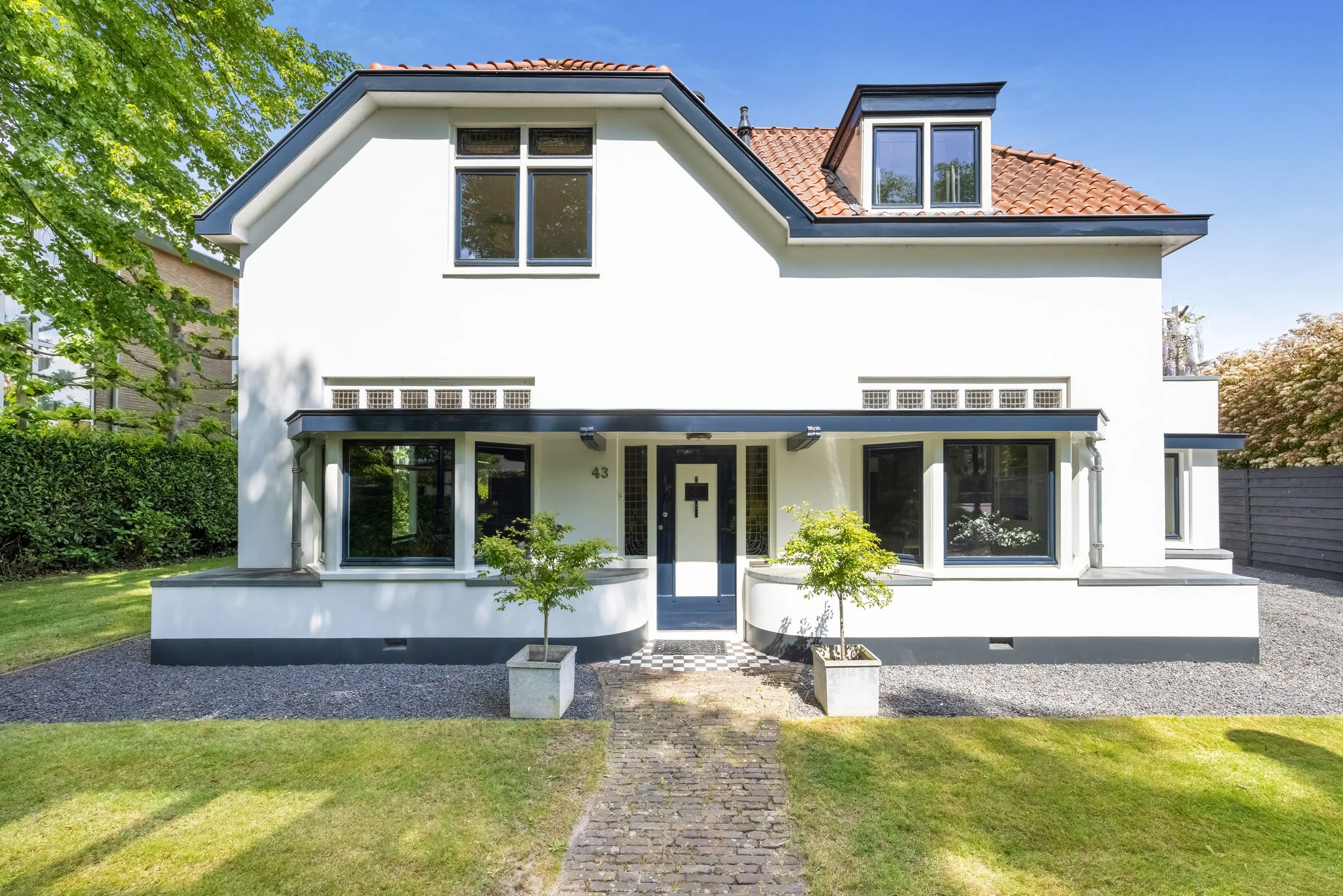 Een witte huis met een rood tegel dak en grote ramen, omgeven door een groene tuin en bomen, onder een blauwe lucht.