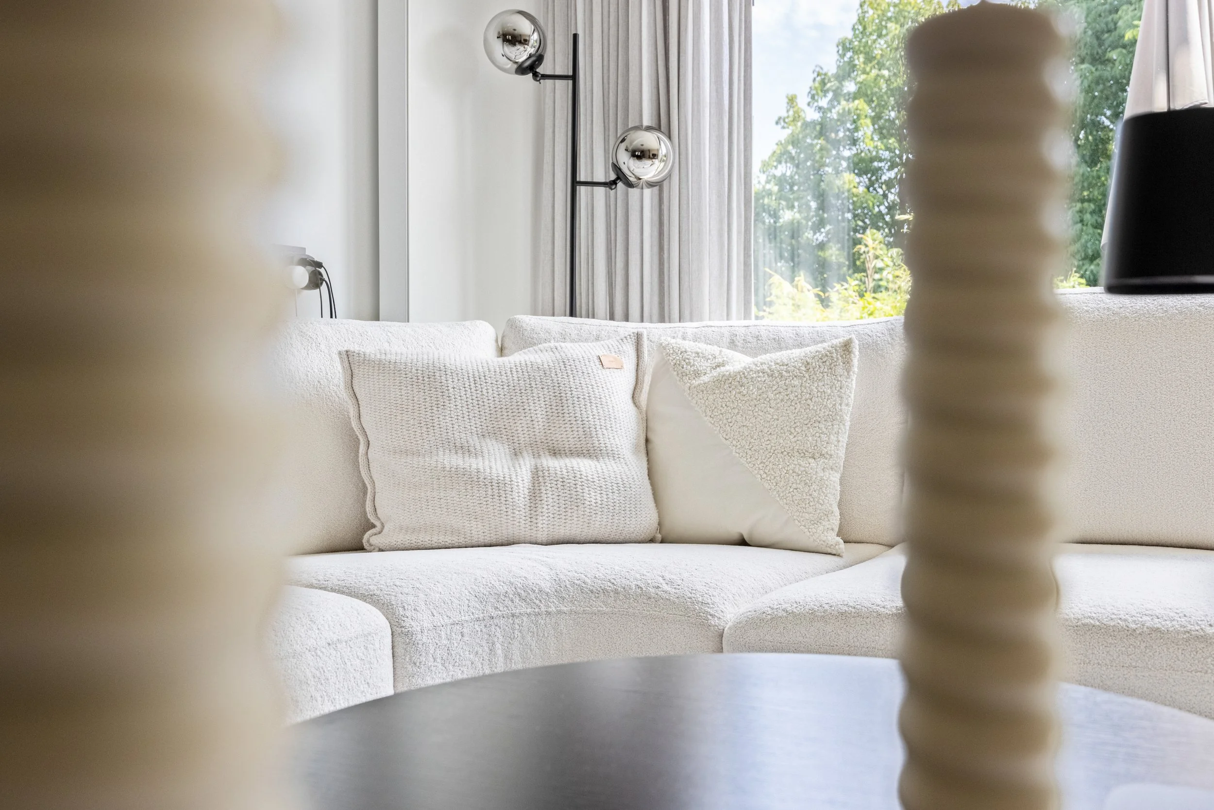 Wit gestoffeerde bank met witte kussens, uitzicht op een raam met groene bomen buiten, zwarte en glazen muurlampen, beige en zwarte decoratieve elementen.