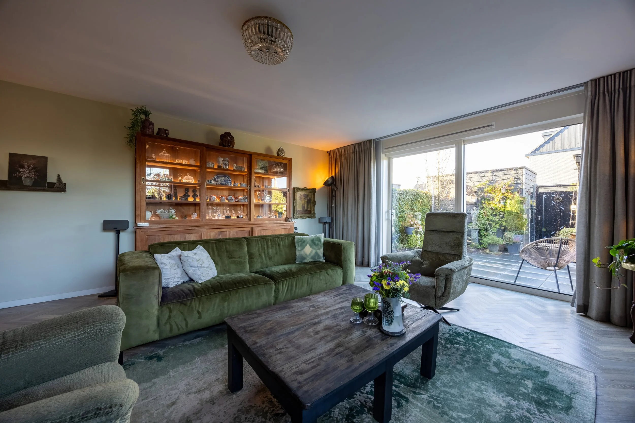 Woonkamer met groene bank, houten kast met glasdeuren, grote ramen met uitzicht op een tuin, bloemen op salontafel, comfortabel eethoek, decoratieve items, gordijnen en vloerbedekking.