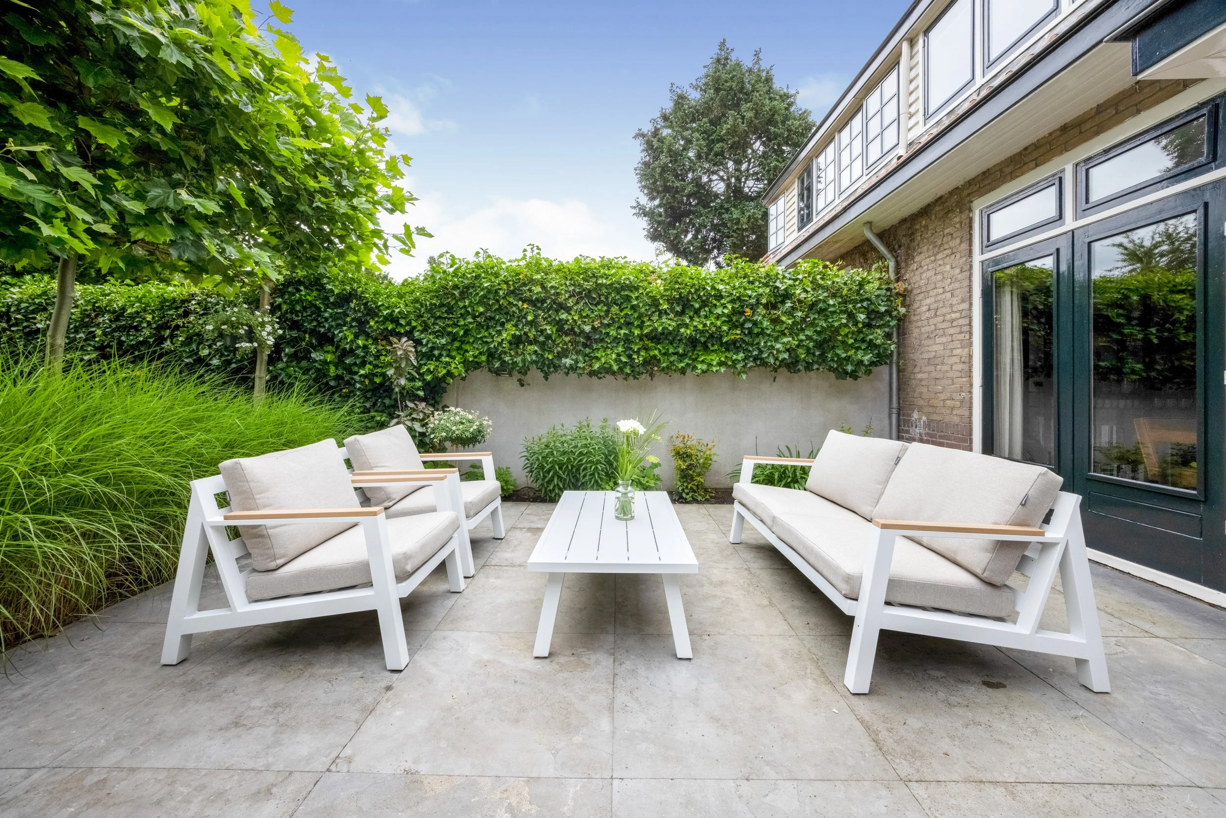 Woonkamer tuin met drie witte banken, een witte tafel met vaas bloemen, groene planten, een boom, en een huis met grote ramen.