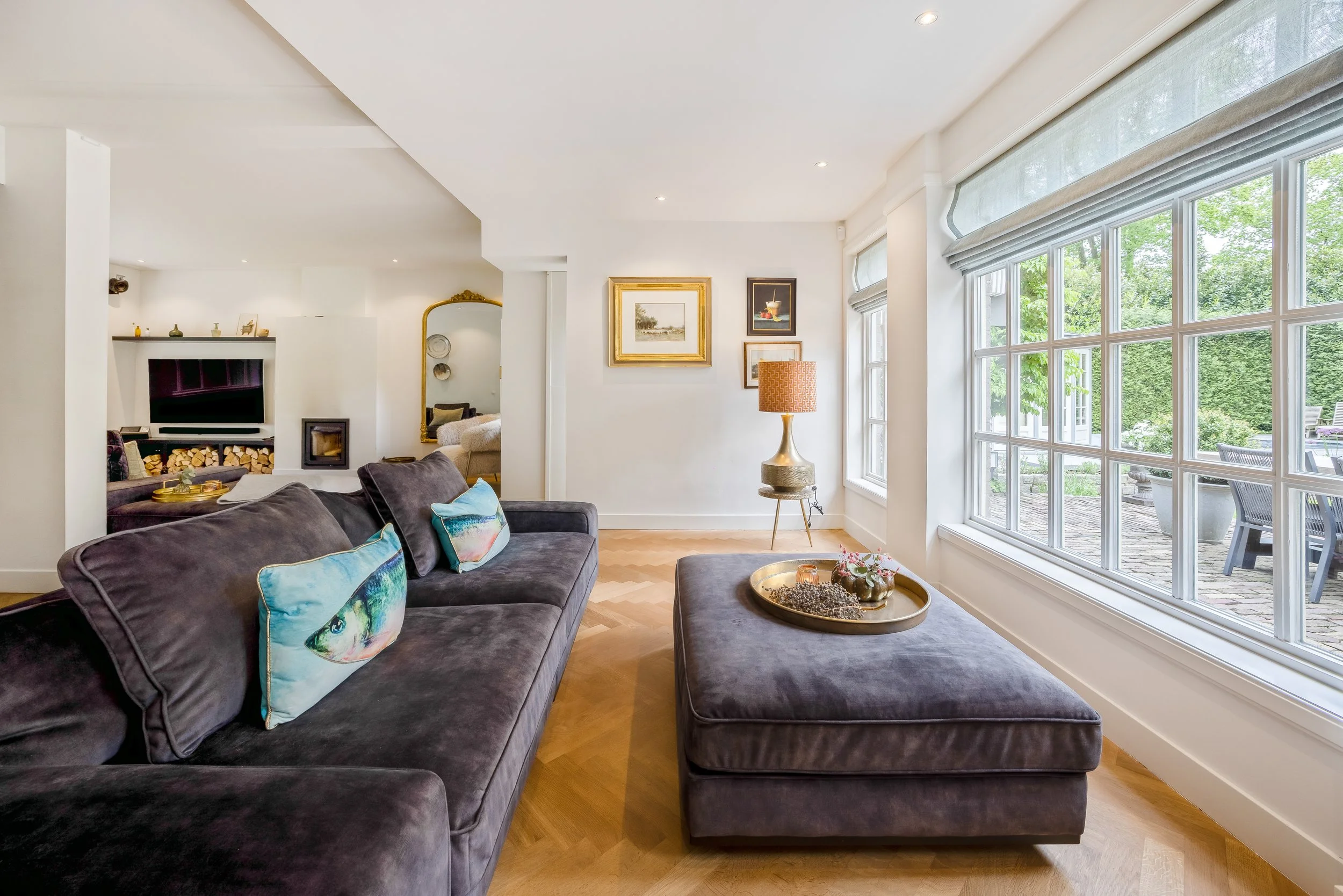 Woonkamer met grote ramen, donkerbruine bank, salontafel met decoraties, muurschilderijen en een grote spiegel.