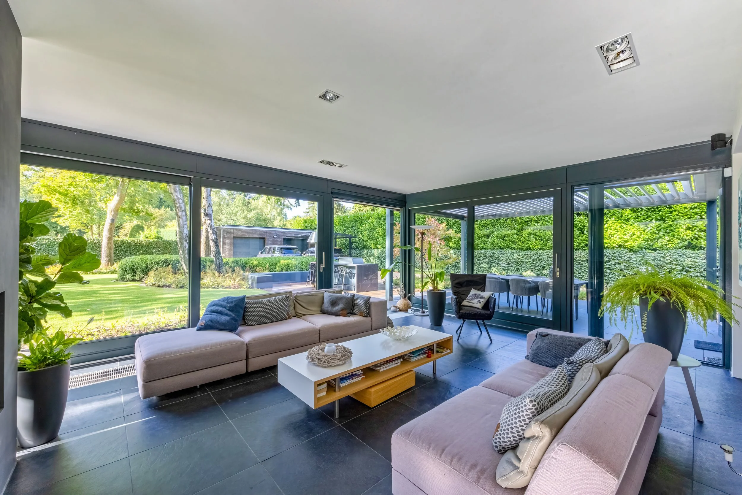 Lichtwoonkamer met grote ramen die uitkijken op de tuin, met roze banken, een witte salontafel, planten en een eettafel buiten.